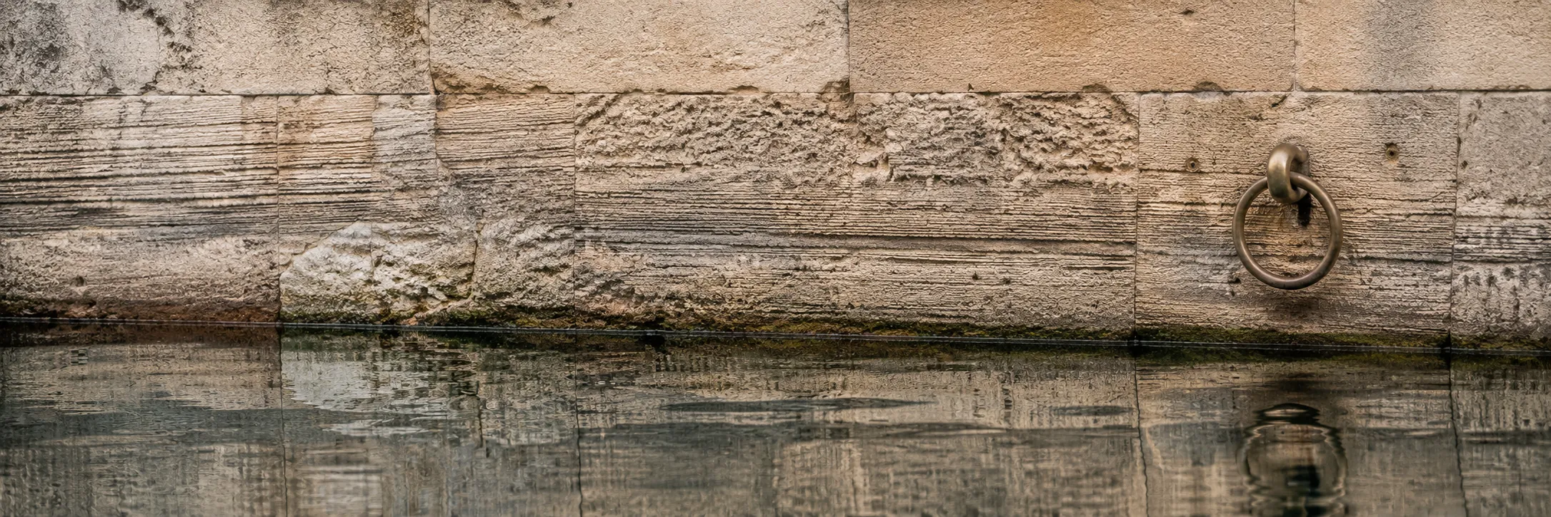 Weathered limestone harbour-wall tide marks with brass mooring ring — money flowing in and out, measured.